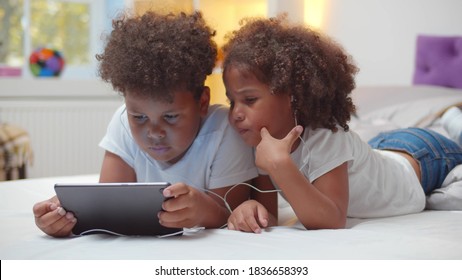 Portrait Of Adorable African Boy And Girl Kids Watching Cartoon In Earphones On Digital Tablet Lying On Bed Together. Brother And Sister Children Sharing Headphones Using Tablet Pc In Bedroom