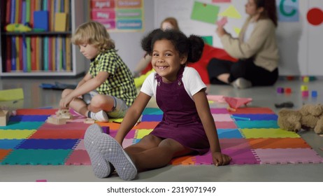 Portrait of adorable African-American preschool girl sitting on floor and smiling at camera in kindergarten. Diverse little kids play in playroom at nursery school - Powered by Shutterstock