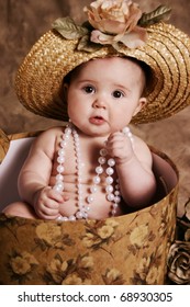 Portrait Of An Adorable Baby Girl Playing Dress Up, Sitting In A Hatbox Wearing A Straw Hat And Pearl Necklace