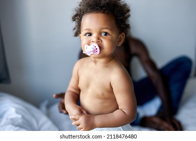 Portrait Of Adorable Black Child Girl With Pacifier In Mouth On Bed At Home, With Dad, Daughter With Dark Skin Look At Camera, Looking Very Charming. Children, Lifestyle Concept