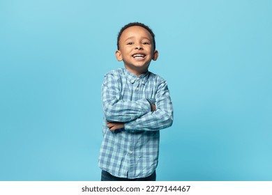 Portrait of adorable black little boy in shirt posing with folded arms and smiling over blue background, copy space. African american male child standing in studio - Powered by Shutterstock