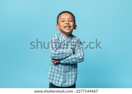 Portrait of adorable black little boy in shirt posing with folded arms and smiling over blue background, copy space. African american male child standing in studio