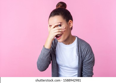Portrait Of Adorable Curious Brunette Teen Girl With Bun Hairstyle In Casual Clothes Peeking Through Fingers, Looking Embarrassed, Pleasantly Surprised. Indoor Studio Shot Isolated On Pink Background