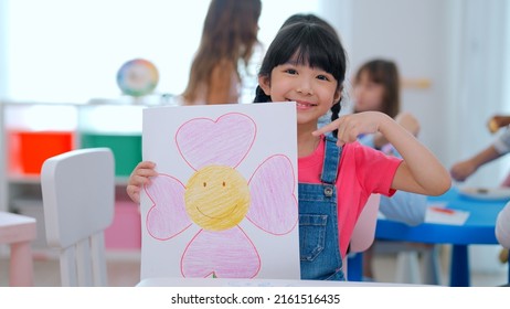 Portrait of adorable girl showing piece of work to camera drawing and painting picture on paper to teacher taking video camera with beautiful smile enjoying in the classroom, Learning with fun concept - Powered by Shutterstock