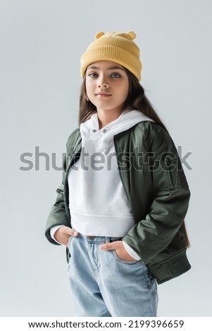 portrait of adorable girl in yellow beanie hat and stylish autumnal outfit posing with hands in pockets isolated on grey