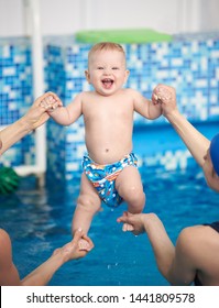 Portrait of adorable happy baby boy with toothy smile dressing in swimming nappies, looking in camera during doing exercising in paddling pool over blue water with cropped female hands helping