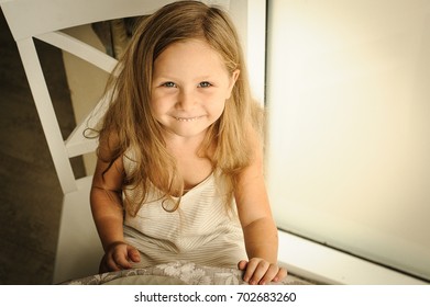 Portrait Of Adorable Happy Young Girl With Long Blonde Spiky Hair. She Sit On White Provence Style Chair And Smile. She Wearing White Striped Dress.Top View, Close Up. White, Beige And Brown Colors.