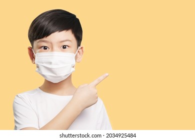 Portrait Of An Adorable Healthy Asian Boy In White T-shirt With Medical Face Mask Looking Wide Eyed To Camera And Pointing Up. Yellow Background, New Normal, Back To School, Poster, Copy Space.