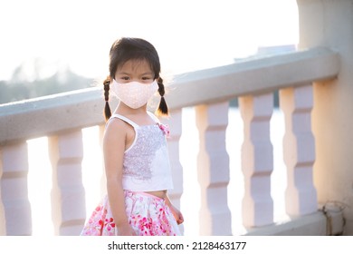 Portrait Adorable Kid Girl Wearing Cloth Face Mask Travel Public Park In Evening. Summer Or Spring Time. Child Stand And Watch Nature At Foot Of Bridge Across Pond. Beautiful Children Aged 5 Years Old