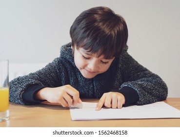 Portrait Of Adorable Kid Sitting Alone Doing Homework With Happy Face, Cute Child Boy Using Rubber Rubbing Wrong Word Written On Paper. Students Doing Spelling Test At Home.Homeschooling Concept