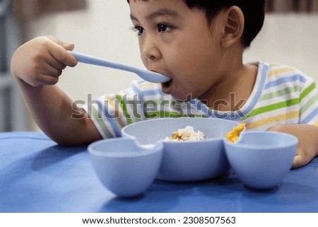 Portrait of adorable little Asian boy eats omelet by using spoon at the table. Kid boy having fun with food at nursery room