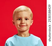 Portrait of adorable little baby boy, blond haired child wearing blue t-shirt and looking at camera with warm smile against red studio background. Concept of childhood, emotions, fashion, lifestyle