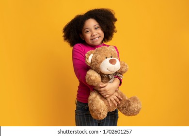 Portrait of adorable little black girl with teddy bear in hands standing over yellow background, posing and smiling to camera, free space - Powered by Shutterstock