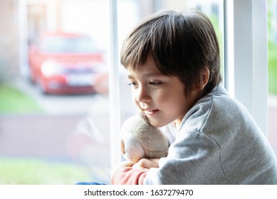 Portrait Adorable Little Boy Sitting Alone Next To Window Looking Out With Smiling Face, Candid Shot Child Playing With Teddy Bear Toy With Blurry Bright Light Background, Kid Relaxing At Home.