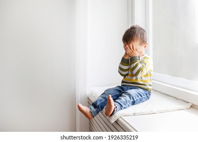 Portrait of adorable little boy sitting on the windowsill and crying. Upset child covering his face at home. Barefoot kid hiding behind palms of his hands. Close up, copy space, background. - Shutterstock ID 1926335219