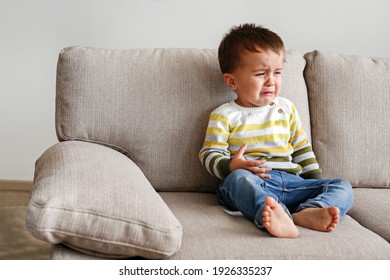 Portrait Of Adorable Little Boy Sitting On The Textile Couch And Crying. Upset Toddler Throwing A Tantrum At Home. Barefoot Kid Calling For Attention. Close Up, Copy Space, Background.