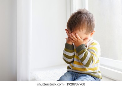 Portrait Of Adorable Little Boy Sitting On The Windowsill And Crying. Upset Child Covering His Face At Home. Barefoot Kid Hiding Behind Palms Of His Hands. Close Up, Copy Space, Background.