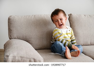 Portrait Of Adorable Little Boy Sitting On The Textile Couch And Crying. Upset Toddler Throwing A Tantrum At Home. Barefoot Kid Calling For Attention. Close Up, Copy Space, Background.