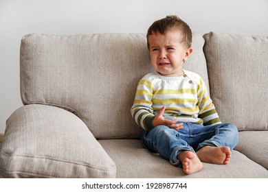 Portrait Of Adorable Little Boy Sitting On The Textile Couch And Crying. Upset Toddler Throwing A Tantrum At Home. Barefoot Kid Calling For Attention. Close Up, Copy Space, Background.