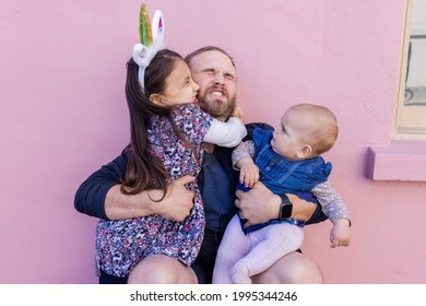 Portrait Of Adorable Little Girl Lovely Hugging Her Father While Holding Cute Baby In Front Of A Pink Wall. Bearded Man With Struggling Facial Expression Holding Cute Daughters. Happy Family Outdoors