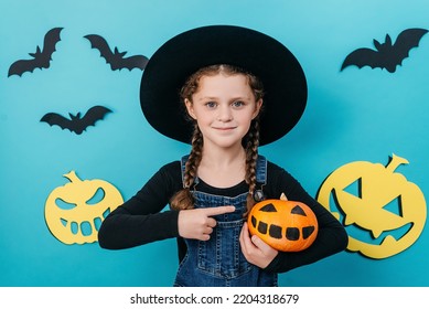 Portrait Of Adorable Little Kid Girl In Black Big Hat Point Finger At Small Funny Pumpkin, Celebrates Halloween Holiday, Posing Isolated On Blue Color Background In Studio With Copy Space For Design