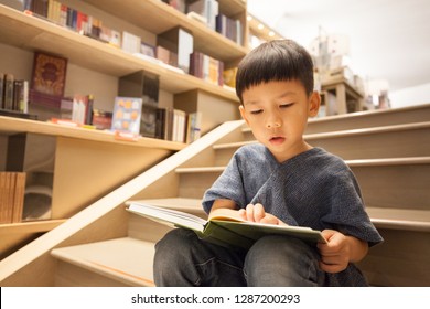 Portrait Of Adorable Little Preschool Asian Boy Sitting On Stairs, Reading Book In Library With Patience, Joy And Concentration. Child’s Brain Development, Learn To Read, Cognitive Skills Concept.