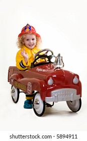 Portrait of an adorable little three year old boy wearing fireman costume sitting in a toy firetruck over white
