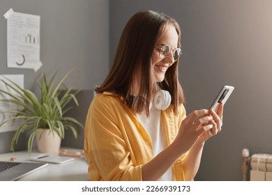 Portrait of adorable positive woman with brown hair wearing yellow shirt and eyeglasses posing in office, holding mobile phone in hands, typing on smart phone. - Shutterstock ID 2266939173