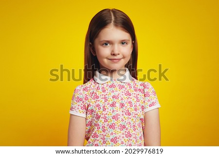 Portrait of adorable schoolgirl in casual summer clothes, looking at camera with happy expression, isolated over yellow background