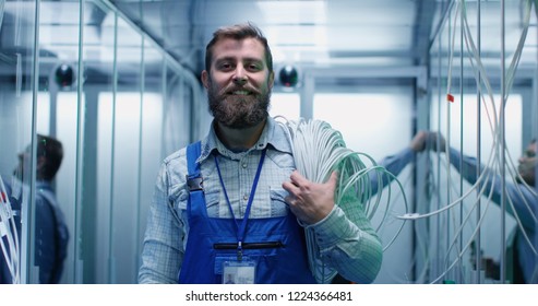 Portrait of adult bearded man technician standing in corridor with cable on his shoulders of server room in data center smiling at camera - Powered by Shutterstock