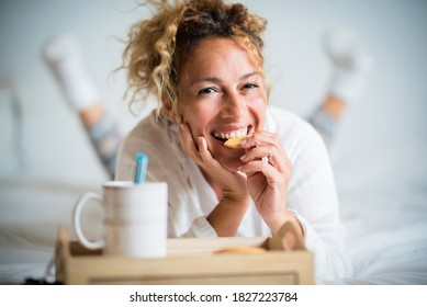 Portrait of adult beautiful woman eating cookie in morning breakfast in the bedroom - home or hotel wake up day with pretty female people lay down with biscuit and tea - cheerful and happy face lady
