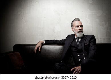 Portrait of adult businessman wearing trendy suit and sitting modern studio on leather sofa against the empty concrete wall. Horizontal, space for ad