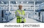 Portrait of Adult Caucasian Male Production Supervisor Looking at Camera And Smiling. Technician in Hardhat And Reflective Jacket at an Industrial Factory with Heavy Machinery Producing Electronics.