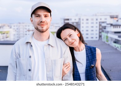 Portrait of adult couple standing on rooftop with city backdrop. Casual clothing, warm expressions, comfort and togetherness. Man wearing cap, woman in plaid outfit with heart-shaped earrings. - Powered by Shutterstock