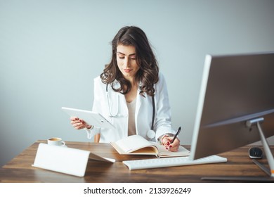 Portrait Of Adult Female Doctor Sitting At Desk In Office Clinic. A Female Doctor Sits At Her Desk And Writes Something On Her Prescription Pad. Portrait Of Female Doctor Wearing White Coat In Office