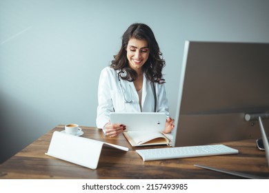 Portrait Of Adult Female Doctor Sitting At Desk In Office Clinic. A Female Doctor Sits At Her Desk And Writes Something On Her Prescription Pad. Portrait Of Female Doctor Wearing White Coat In Office