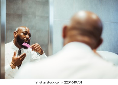 A Portrait Of An Adult Handsome Bearded Bald Black Man Entrepreneur In A White Shirt With A Necktie Combing His Beard With A Pink Comb In A Public Restroom In Front Of The Mirror