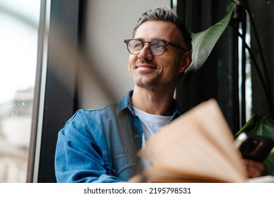 Portrait of adult handsome smiling man in glasses looking out the window while sitting with book in cafe - Powered by Shutterstock