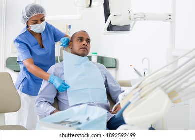 Portrait Of Adult Hispanic Man Sitting On Dental Chair While Woman Dentist Preparing Him For Medical Examination In Clinic