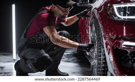Portrait of an Adult Man Working in a Detailing Studio, Prepping a Factory Fresh American Sportscar for Maintenance Work and Car Care Treatment. Cleaning Technician Using Sponge to Wash the Wheels