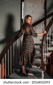 Portrait Adult Woman Actress Posing Dressed Retro Styling Clothes On Home Staircase In Vintage Living Room. Female In Old Style Color Dress In Flowers. Concept Retro Fashion And Outdated Trend Vogue