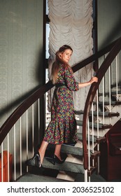 Portrait Adult Woman Actress Posing Dressed Retro Styling Clothes On Home Staircase In Vintage Living Room. Female In Old Style Color Dress In Flowers. Concept Retro Fashion And Outdated Trend Vogue