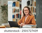 Portrait of adult woman as female teacher sitting at desk and smiling at camera in school library setting