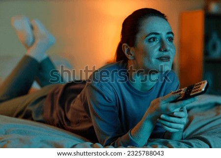 Portrait of adult woman lying on bed at home and watching TV in dark