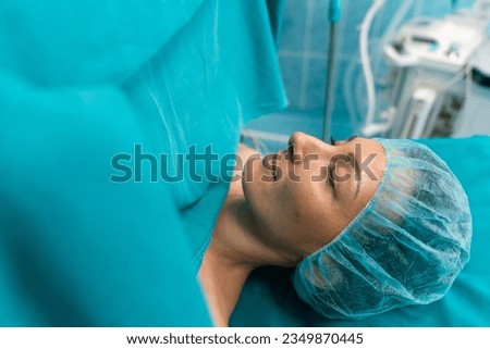 Portrait of adult woman patient with eyes closed sleeping under anesthesia with medical sheets on during surgery in operating room.