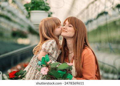 Portrait of affectionate adorable small botanist in floral dress standing at greenhouse and kissing her mother in a cheek. Loving family sharing precious moments at greenhouse. Mother and daughter