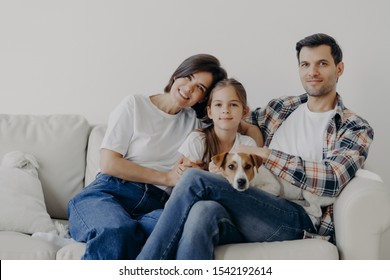 Portrait Of Affectionate Family Cuddle And Sit Together At Couch In Living Room, Change Their Home, Have Happy Expressions. Father, Mother, Daughter And Dog Pose For Making Portrait, Spend Good Time