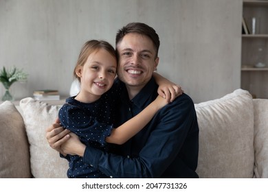 Portrait Of Affectionate Happy Young Hispanic Dad Cuddling Adorable Small Kid Daughter, Showing Sweet Tender Feelings, Sitting On Sofa. Joyful Bonding Two Generations Family Looking At Camera.