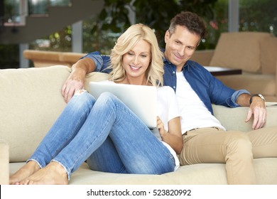 Portrait Of An Affectionate Middle Aged Couple Sitting With A Laptop At Home. Beautiful Blond Woman Using Laptop While Handsome Man Sitting Next To Her And Enjoy Surfing On Internet.