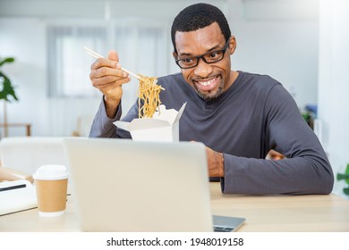 Portrait Of Afican Business Man Enjoying Chinese Food In Office Lunch Break Online Conference, Employee Holding Chopsticks Eating Takeaway Noodle Box Meal Virtual At Workplace In Home Office. 